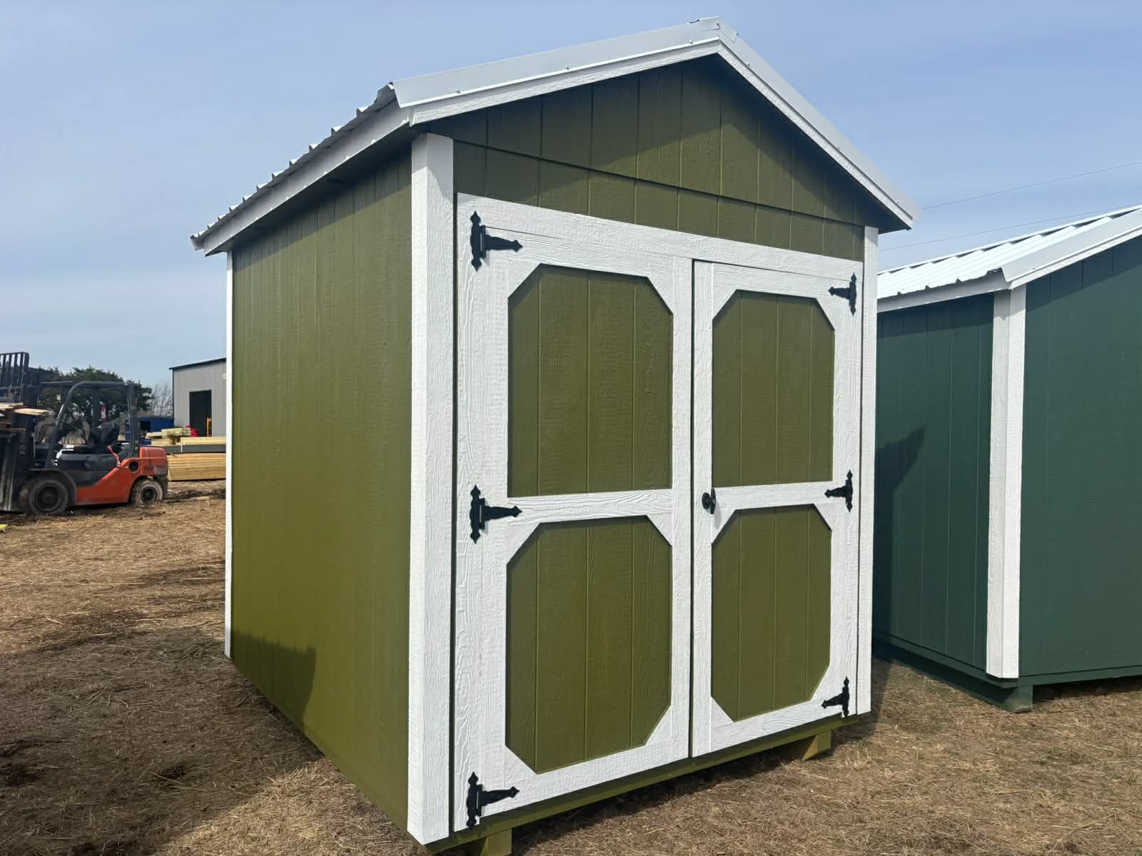 An olive-green shed with white trim and double doors, standing in an outdoor lot next to another similar structure.