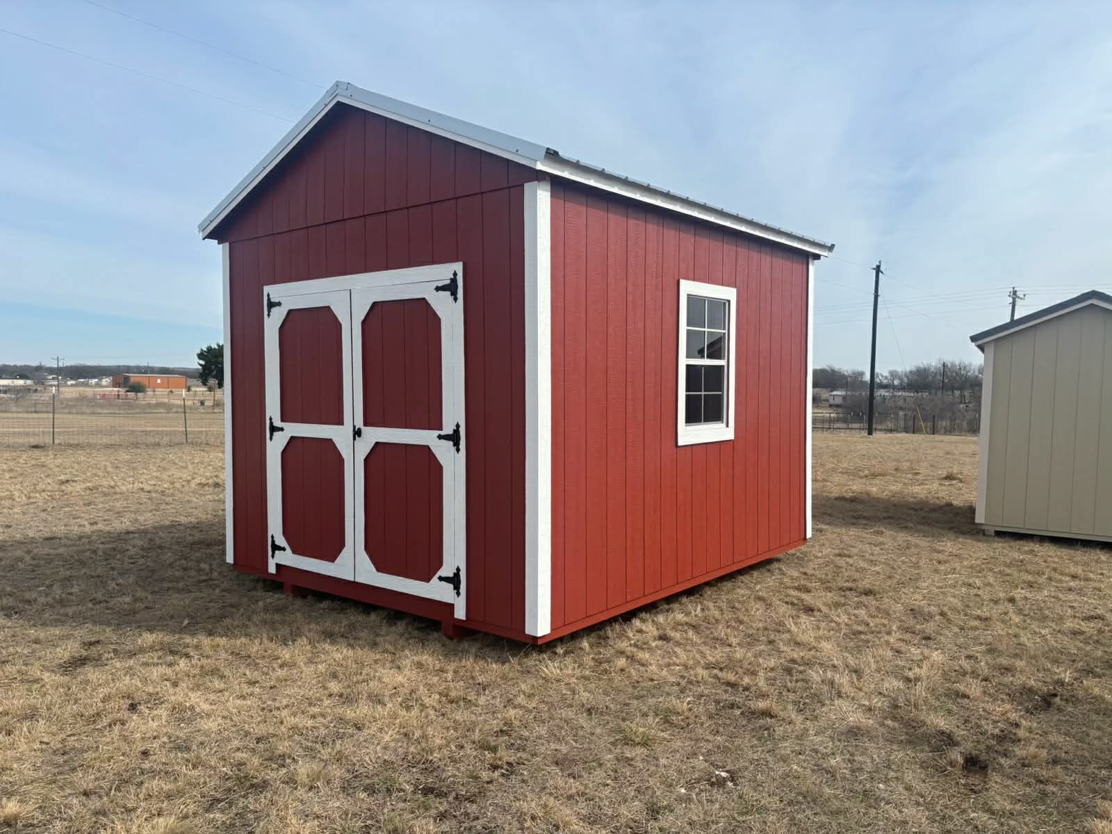 A red wooden shed with white trim, a double door, and a single window sits in a grassy, open field under a blue sky.