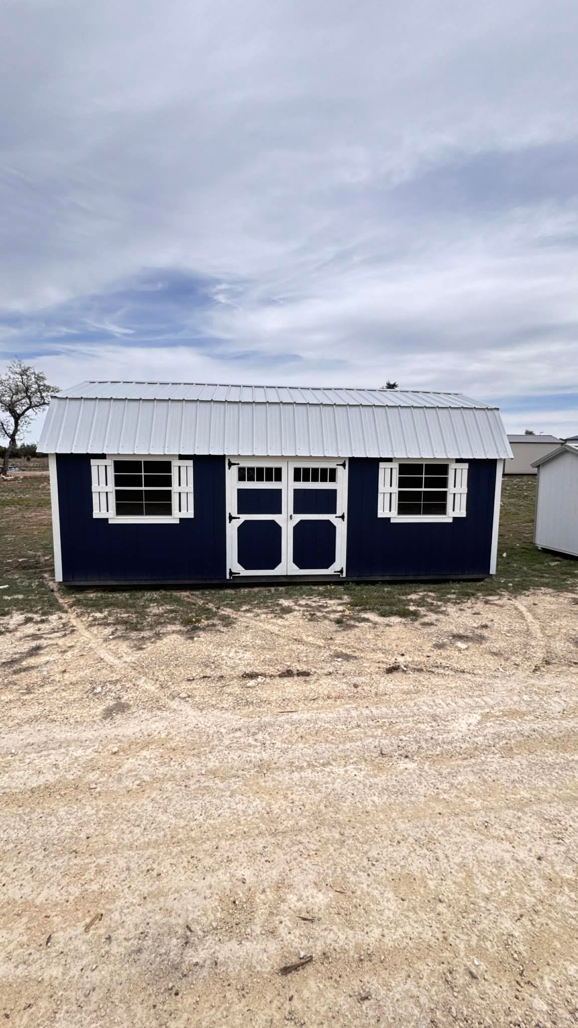 A navy blue storage shed with white trim and a metal roof, positioned on a gravel lot under a cloudy sky.