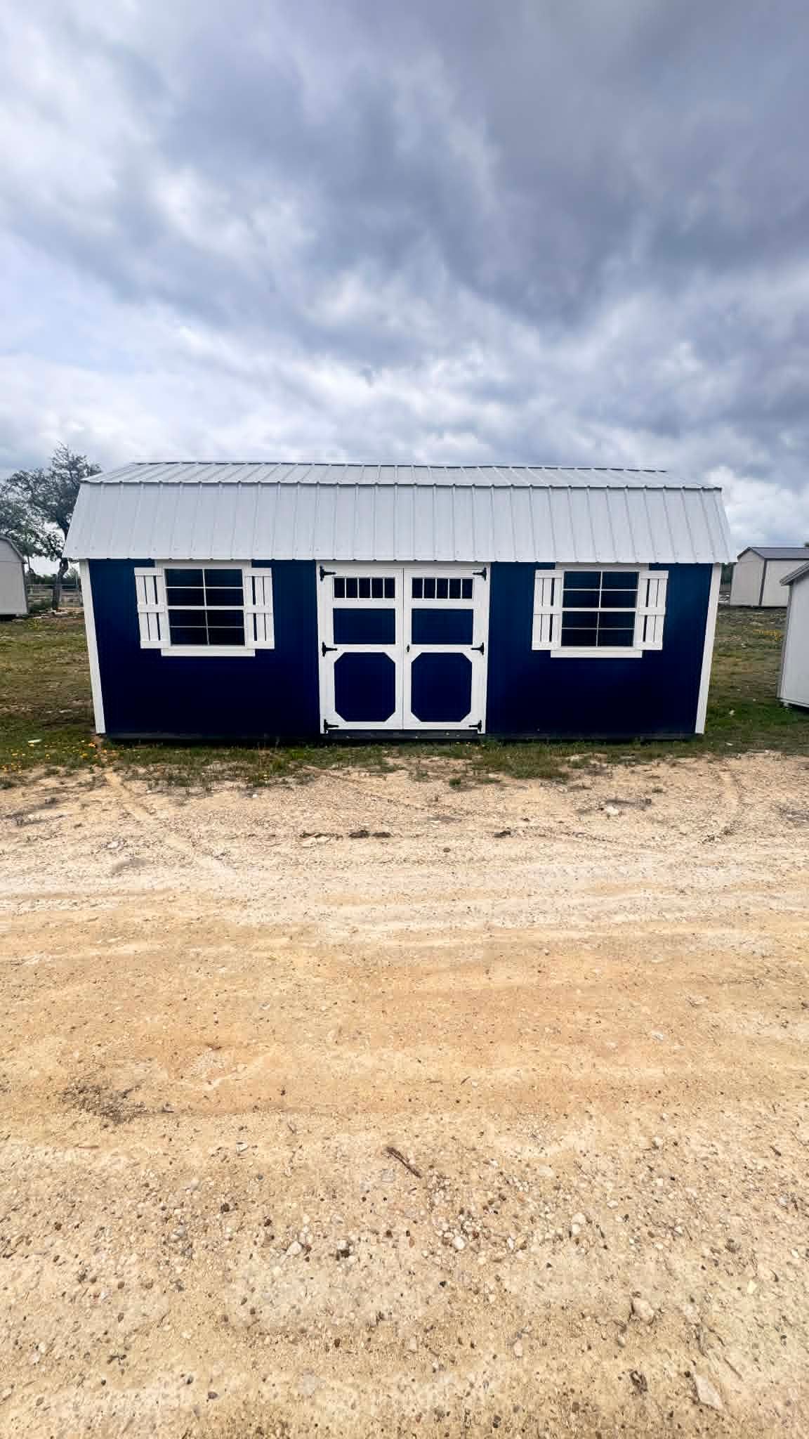 Small blue building with white trim and a metal roof on a gravel lot under cloudy sky