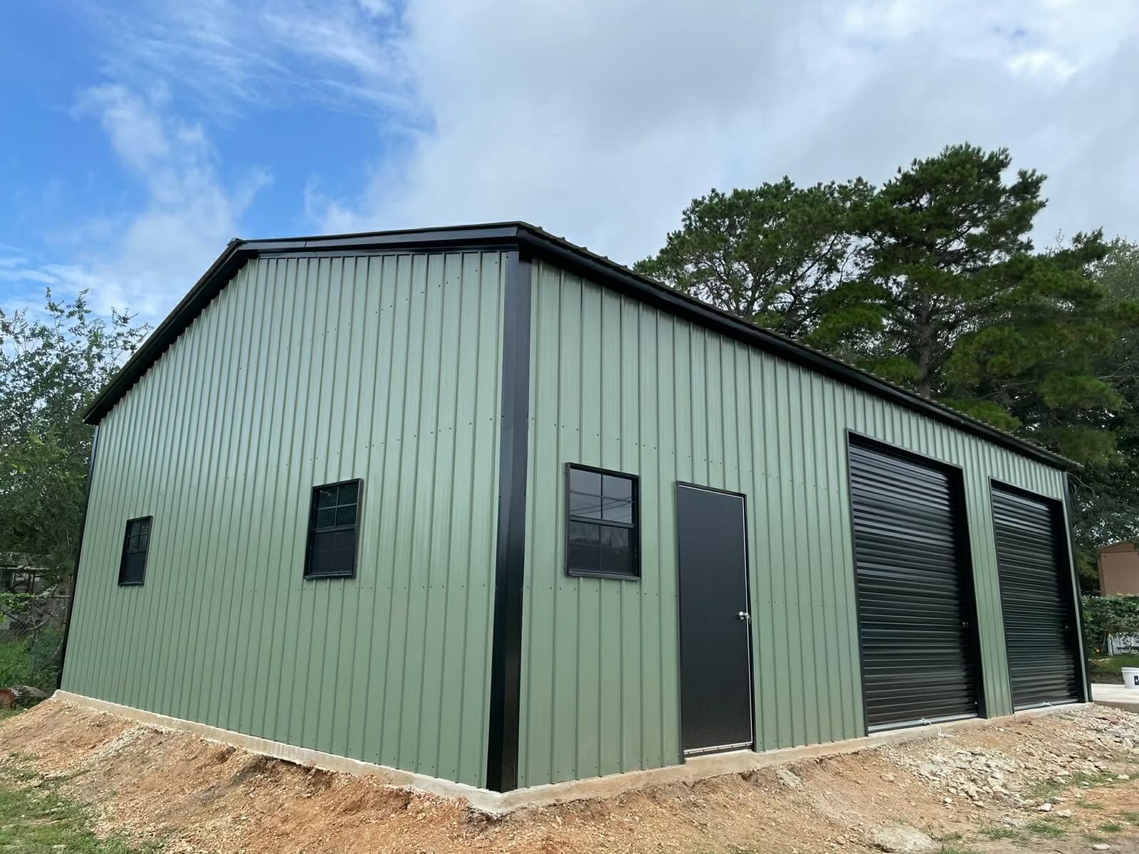 Green metal storage building with black doors and windows on a gravel lot under a cloudy sky