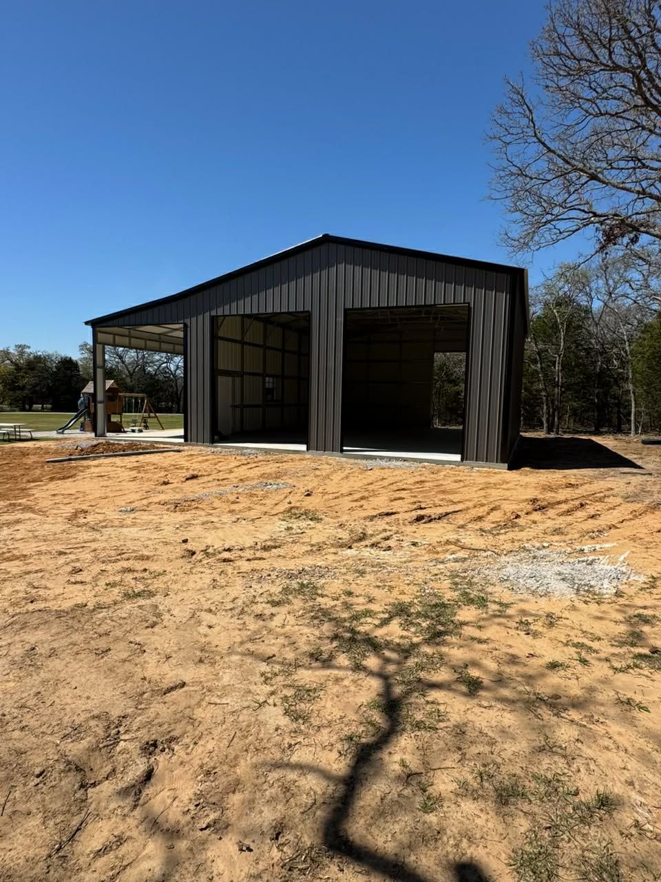 Black metal barn-style workshop with open bays on a dry dirt lot under a clear blue sky