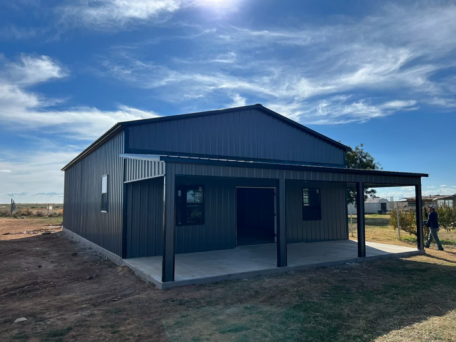 Dark metal shed with porch in a rural dirt lot under a cloudy blue sky