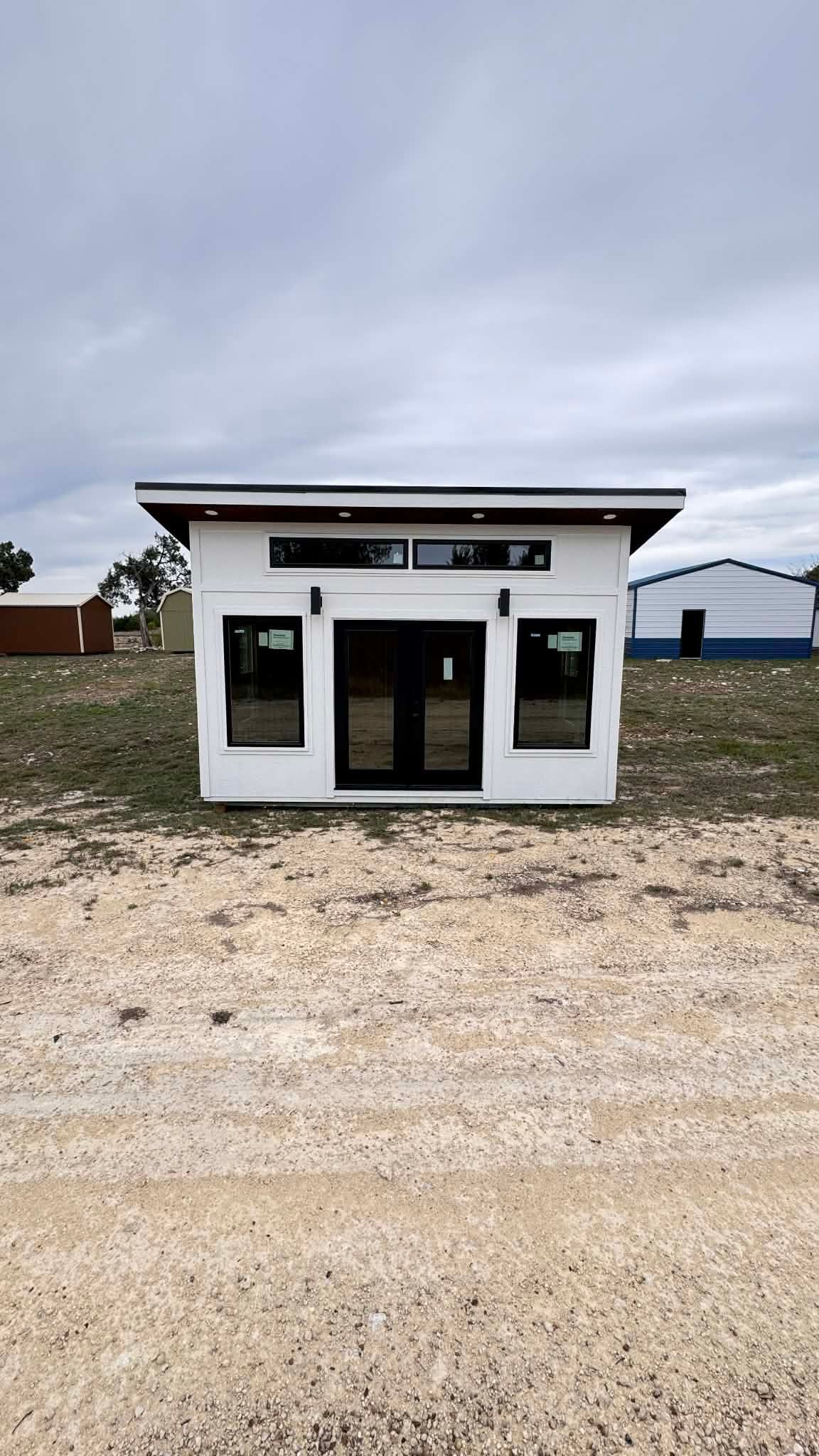 A modern, white, rectangular cabin with a dark roof and black doors and windows stands on a gravel lot under a cloudy sky.