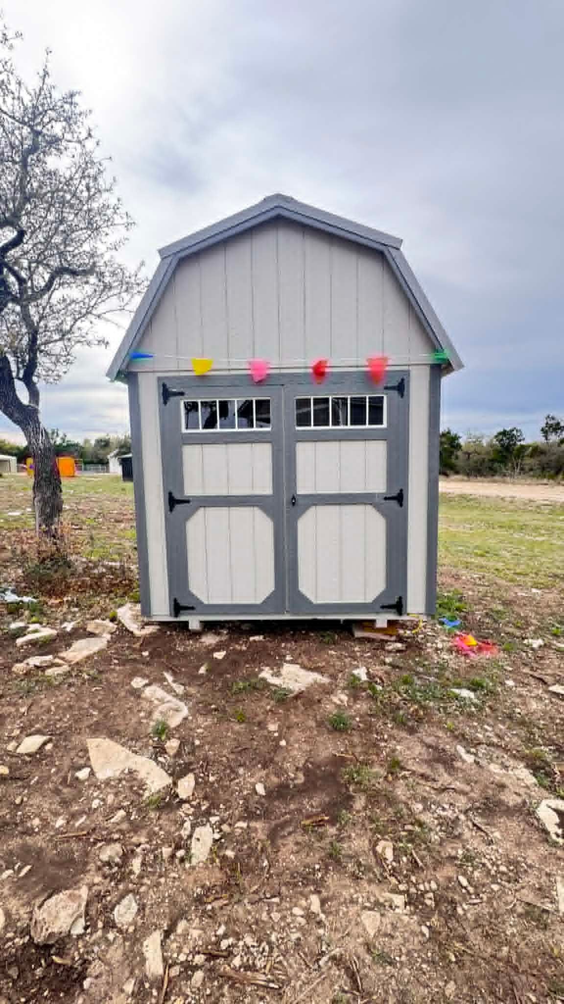 Small gray shed with double doors and colorful bunting, standing on a dirt lot under a cloudy sky.