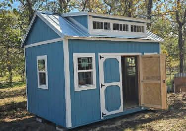 A blue wooden shed with white trim, a metal roof, and a clerestory window section, situated in a wooded area.