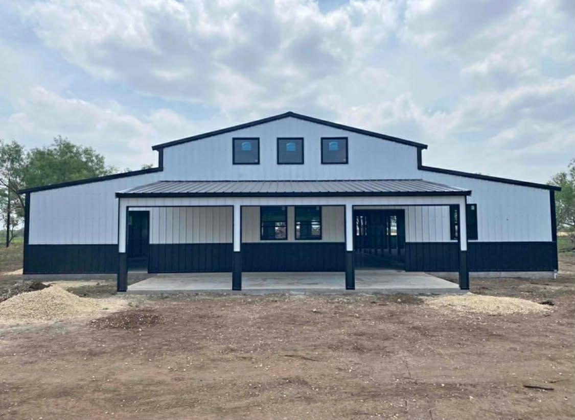 A modern white and black metal barn with a covered porch and three small windows under the roof peak against a blue sky.