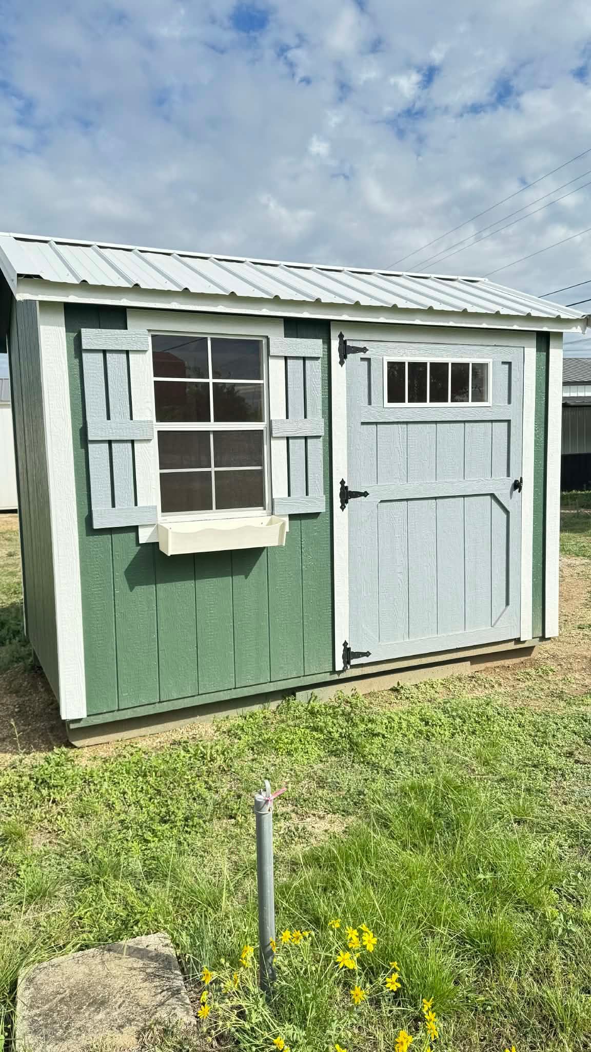 Small green shed with white trim, a window, and a light blue door in a grassy yard under a cloudy sky