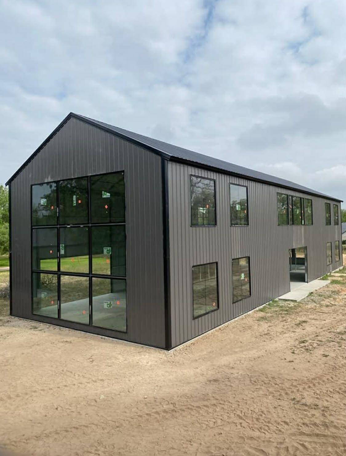 A newly constructed dark grey metal barn-style building with a large glass wall and multiple windows on a dirt lot.