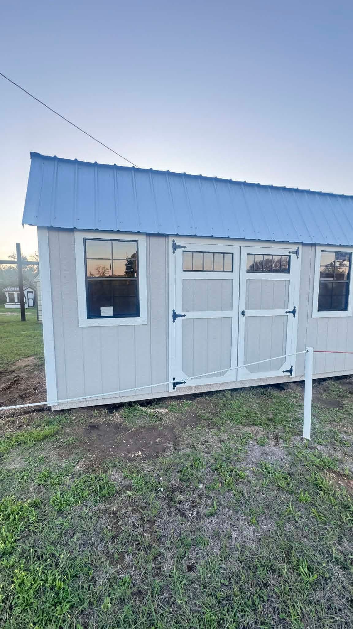 Small white shed with blue metal roof and screened doors in a grassy yard at dusk