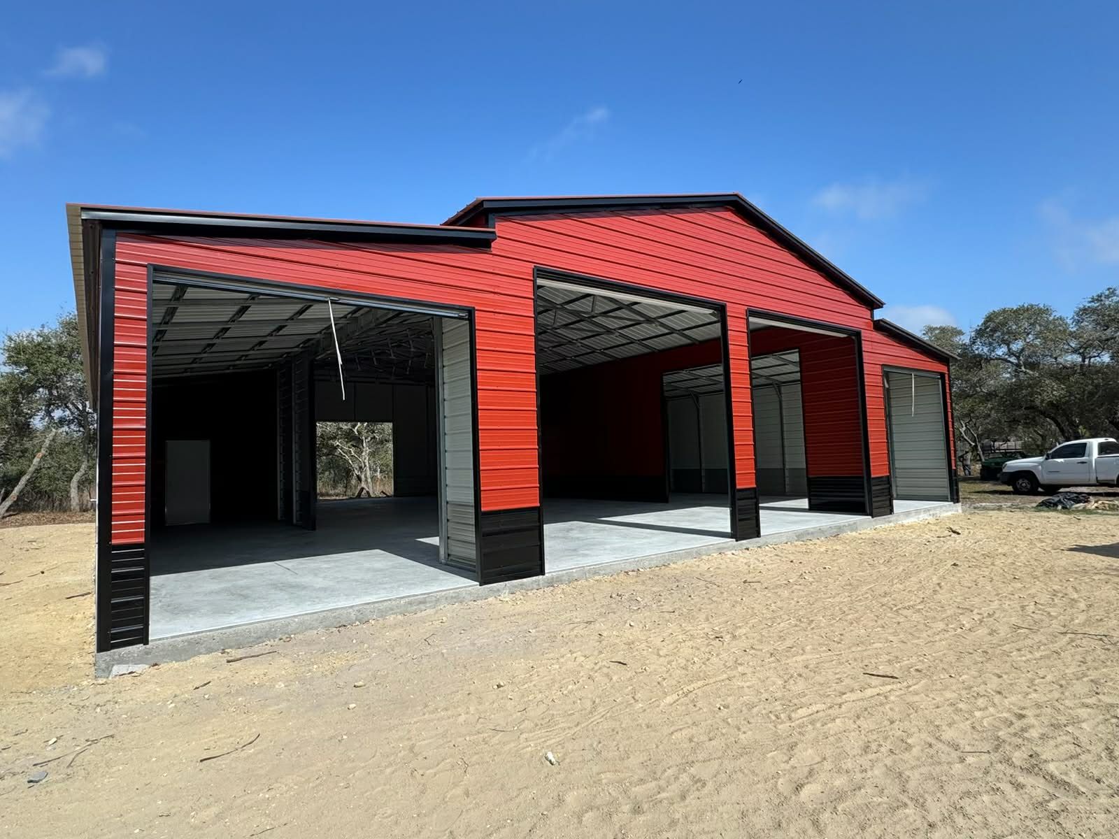 A large, modern, dark gray metal barn-style building with a two-story glass facade, set on a sandy construction site.