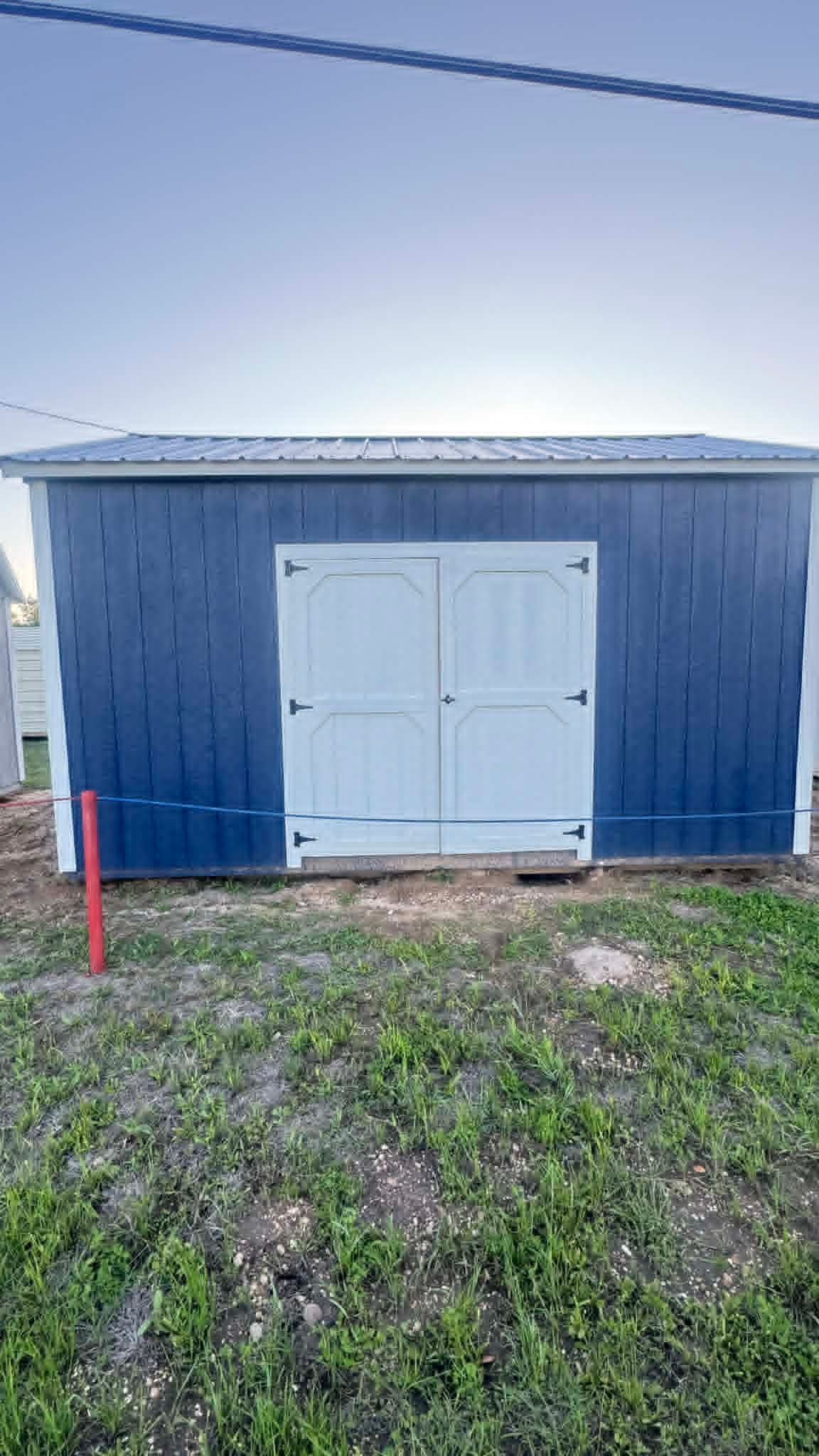 Small blue shed with white double doors on a grassy lot at dusk