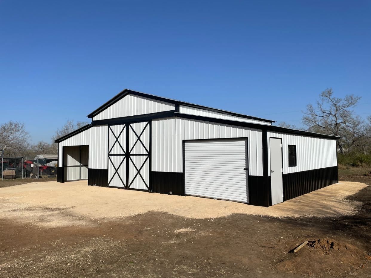 White metal barn-style building with black trim and roll-up doors on a dirt lot under a blue sky