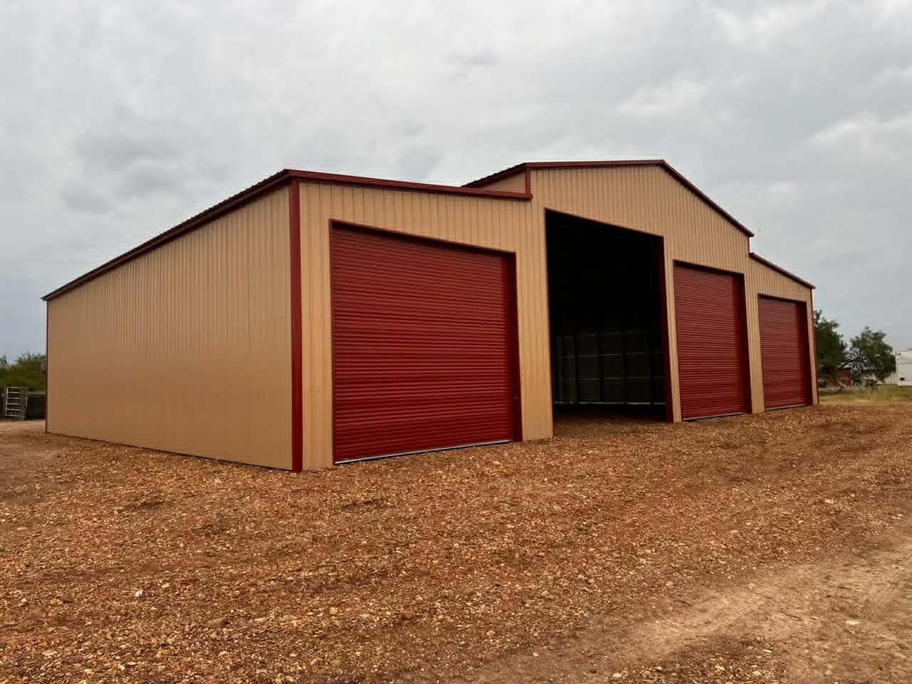 Tan metal storage shed with red roll-up doors on a gravel lot under an overcast sky
