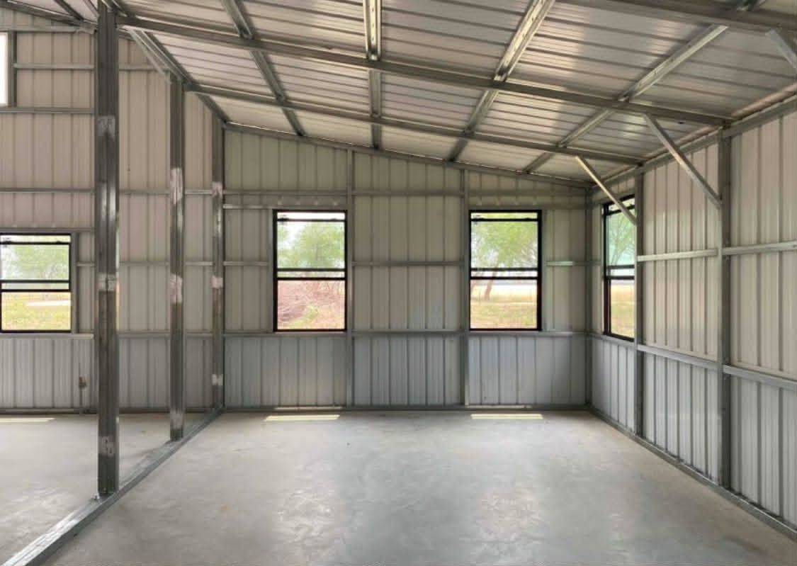Empty metal workshop interior with concrete floor, corrugated walls, and several windows.