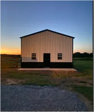 Small beige barn with black trim at dusk, centered on a gravel and grassy farmyard.