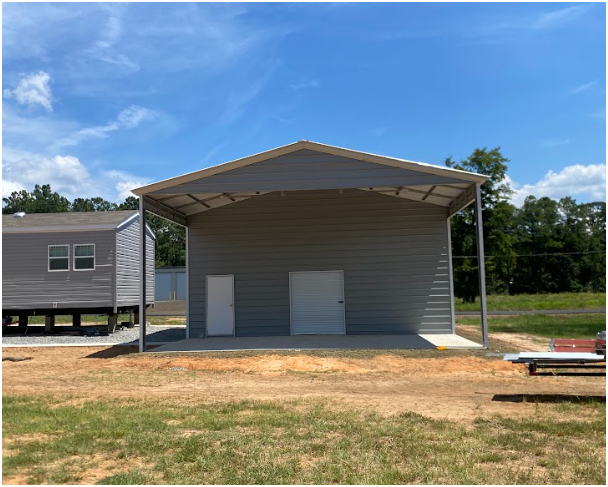 A gray metal building with a white walk-in door and garage door under a covered porch, set on a dirt lot near a house.