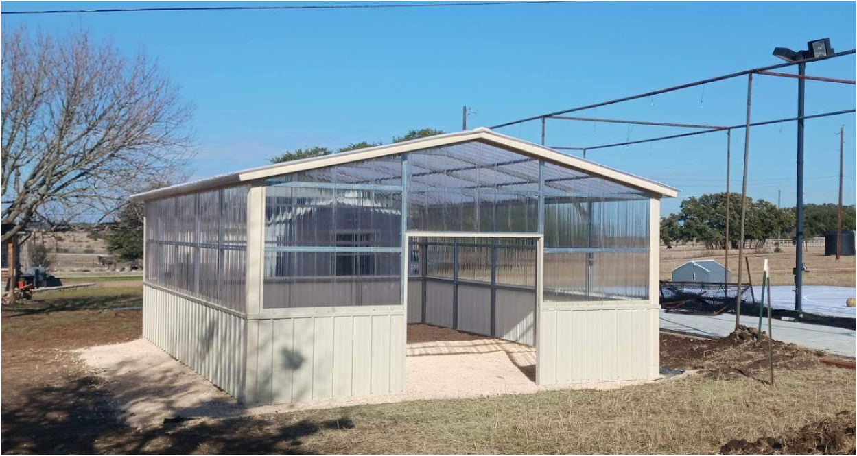 A rectangular greenhouse with beige metal siding and a translucent roof, situated in an open field under a blue sky.