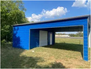 Blue outdoor shed with an open carport-style bay on a grassy lot under a partly cloudy sky