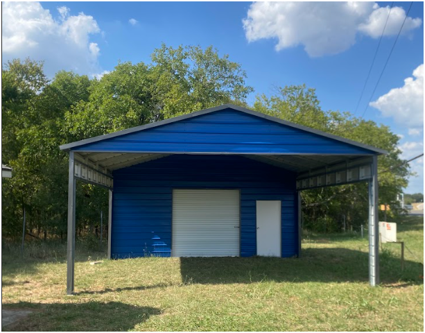 A blue metal storage shed with a carport, a white roll-up door, and a white side door in a grassy area with trees.