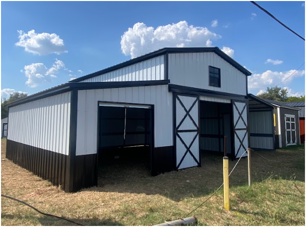 A white and black metal barn with open bays sits on a grassy lot under a blue sky with white clouds.