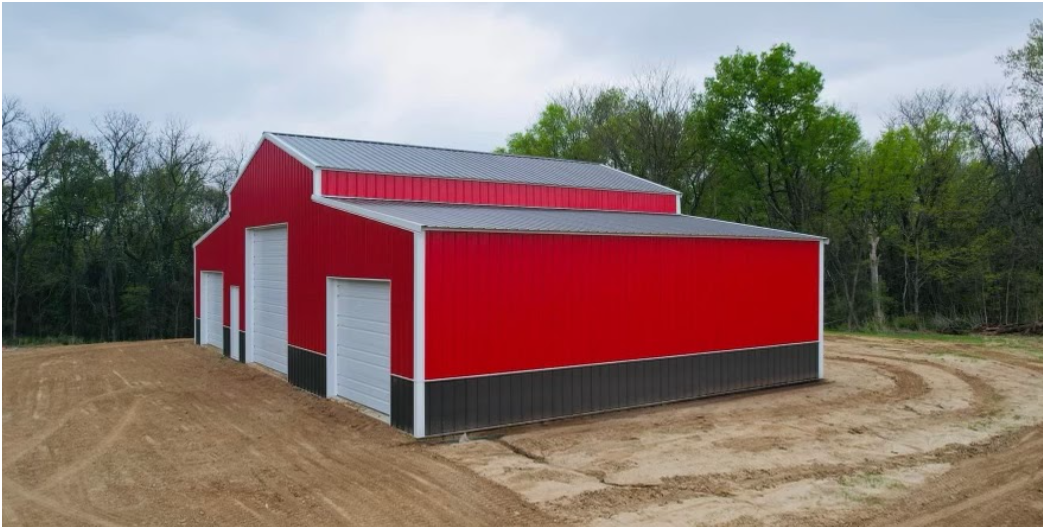 A red and dark gray metal barn with white trim and garage doors stands on a dirt lot near a line of trees.