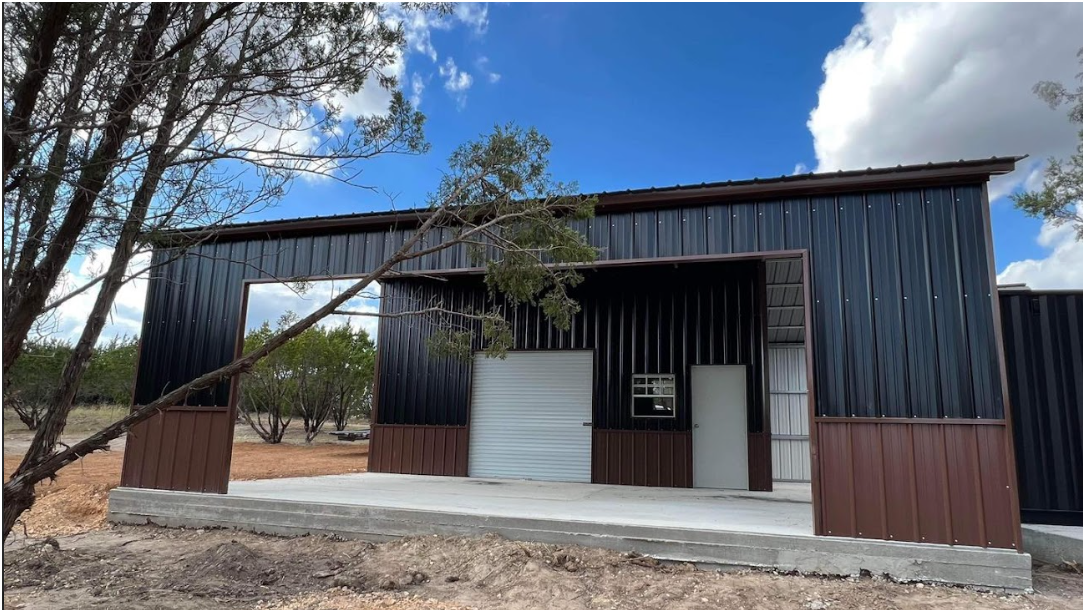 A metal-sided building with black and rust-colored panels, a concrete foundation, a white door, and a partially open bay.