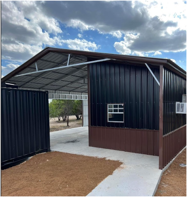 A black metal building with a large open-sided carport, a single window, and a concrete floor under a cloudy sky.