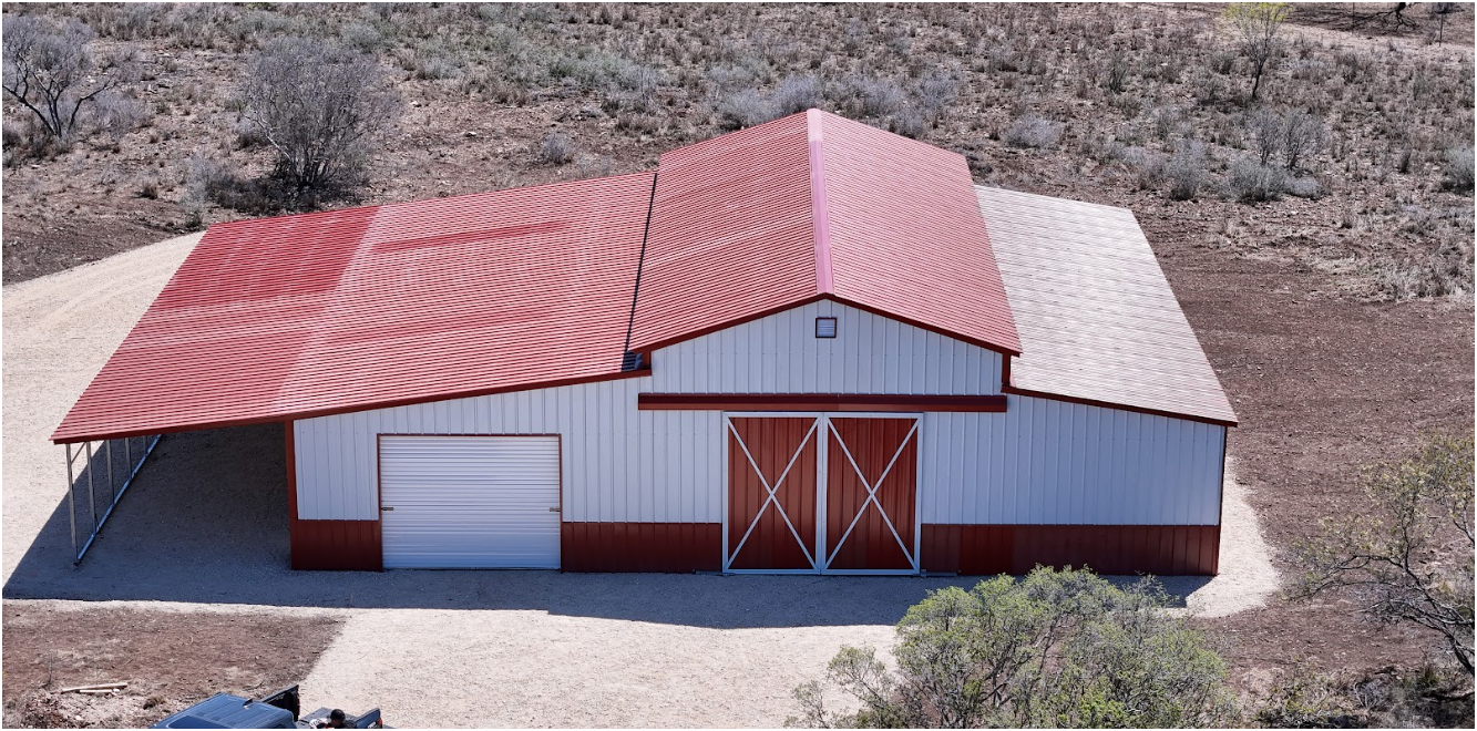 An aerial view of a white, metal-sided barn with a red roof, a roll-up door, and large double doors in a rural landscape.