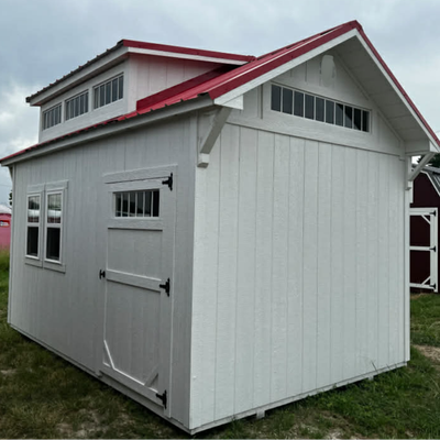 A white storage shed with a red metal roof, a side door, multiple windows, and a raised central roof section.