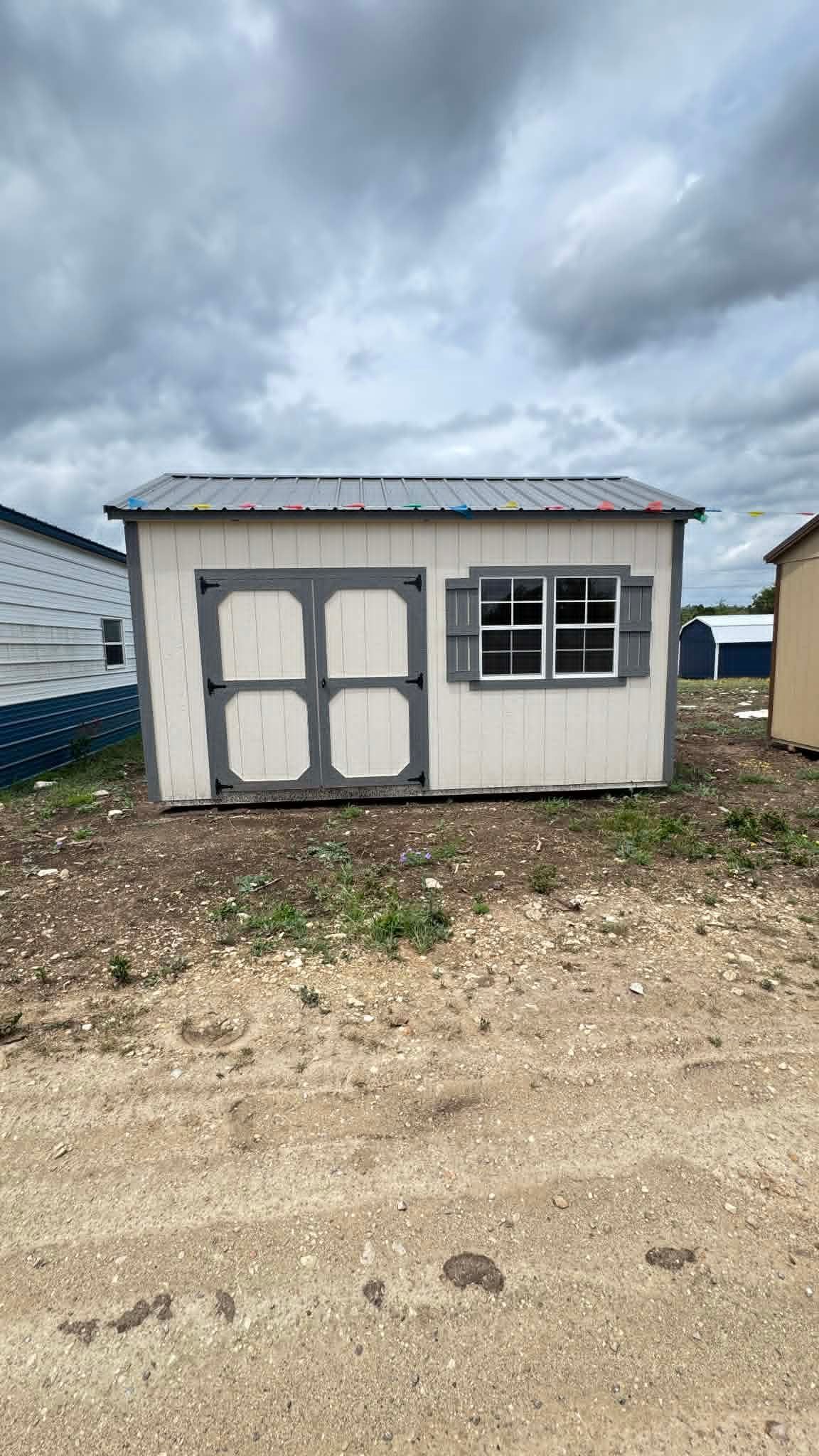 Small white storage shed with gray double doors and a window, under a cloudy sky on a gravel lot