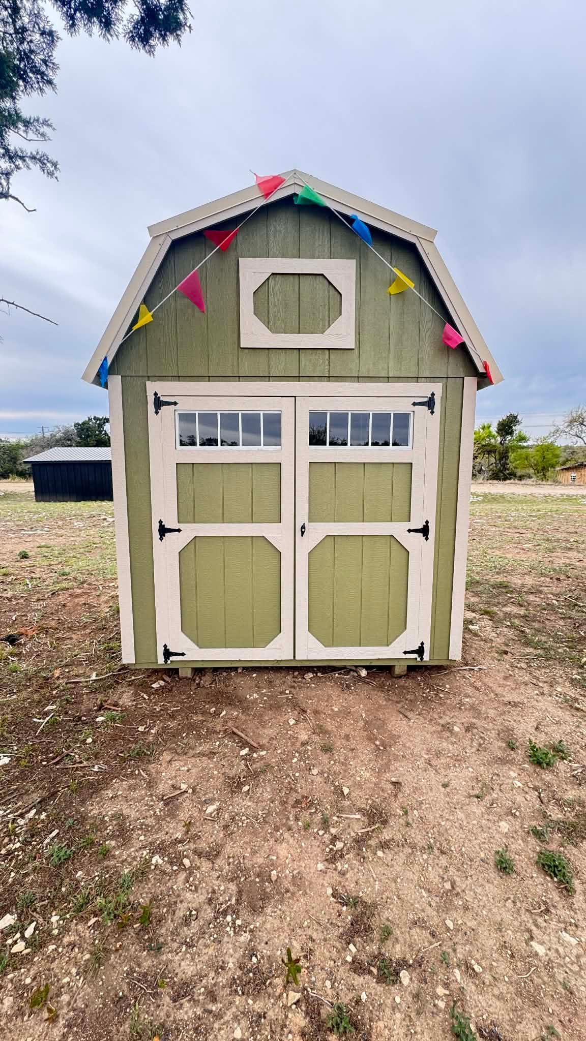 A sage green wooden storage shed with double doors and a gambrel roof, decorated with colorful pennant flags outdoors.