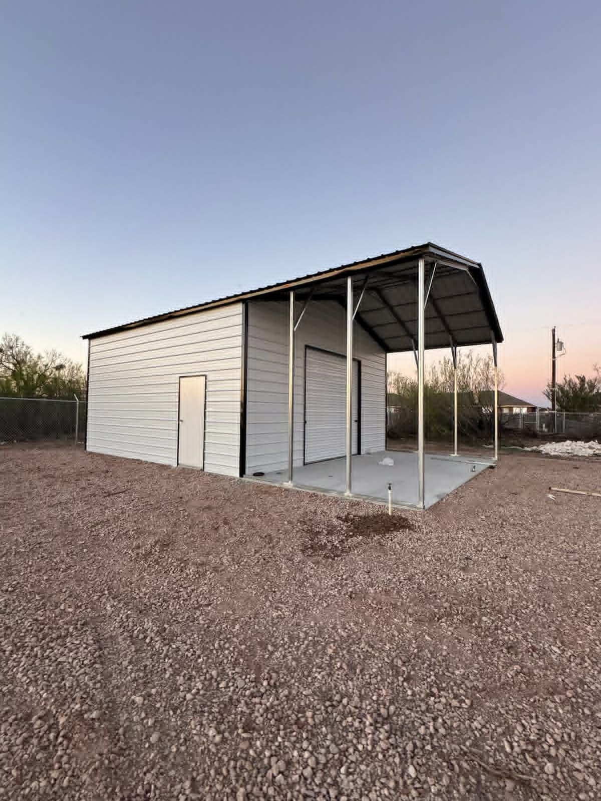 Small white metal shed with a covered porch on a gravel lot at sunset.
