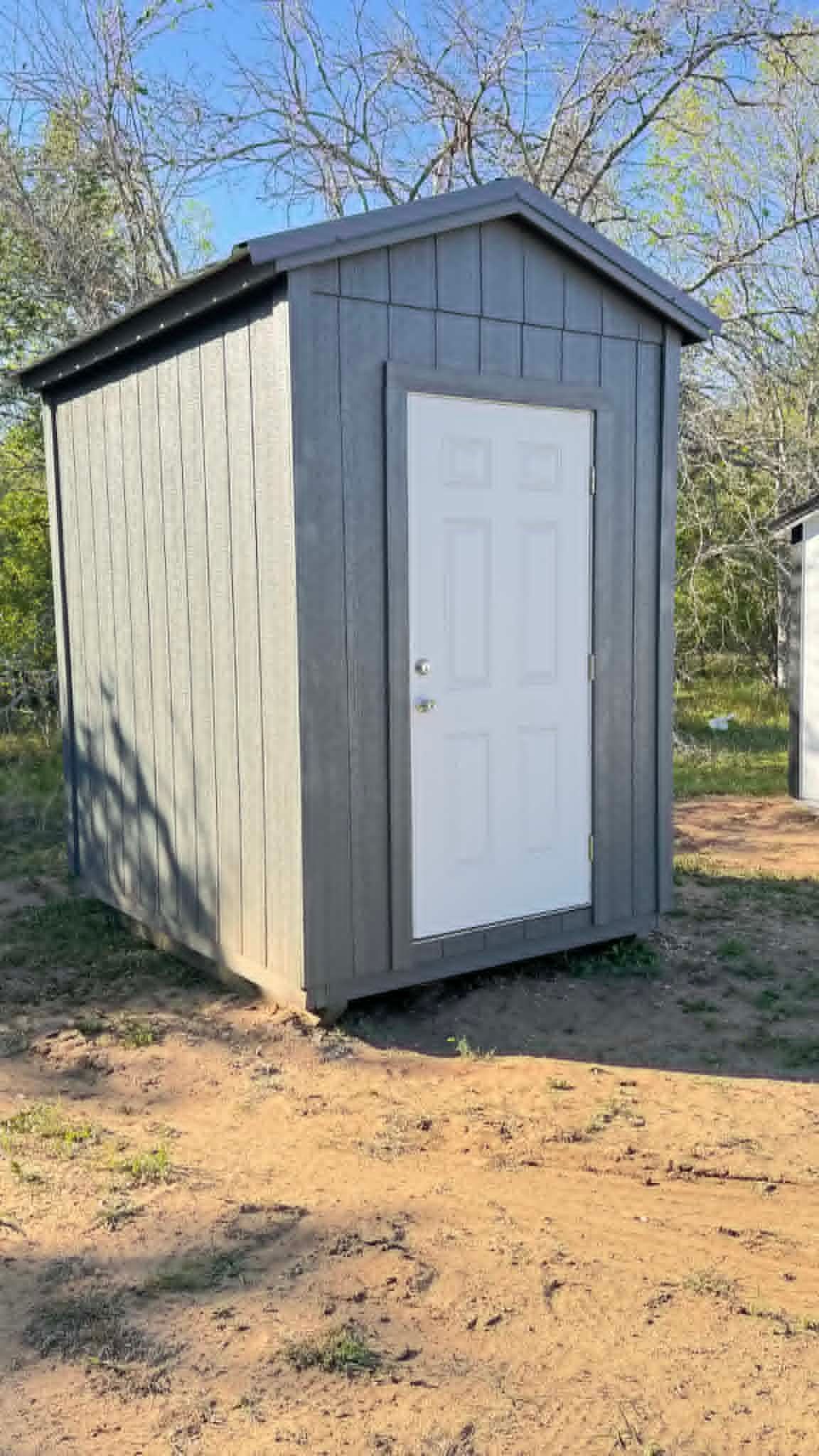 Small gray shed with white door standing on bare dirt outdoors among leafless trees
