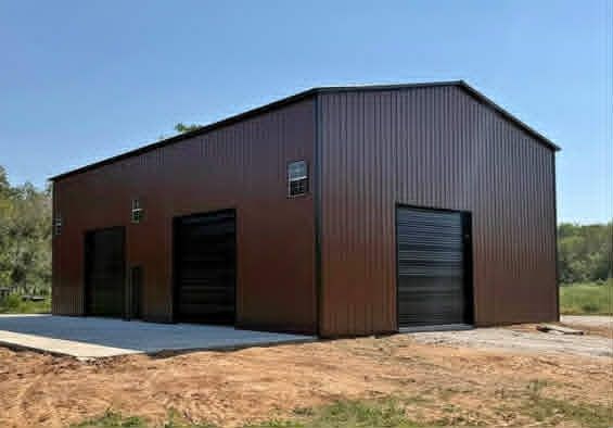 Large brown metal storage building with three garage doors on a concrete pad, set in a rural lot.