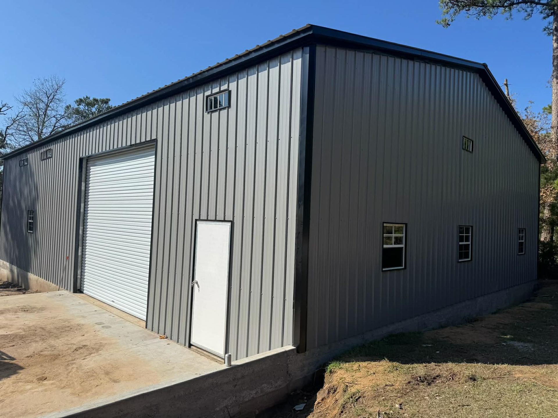 A gray metal warehouse building with a large rolling door, a white walk-in door, and several windows on a dirt lot.