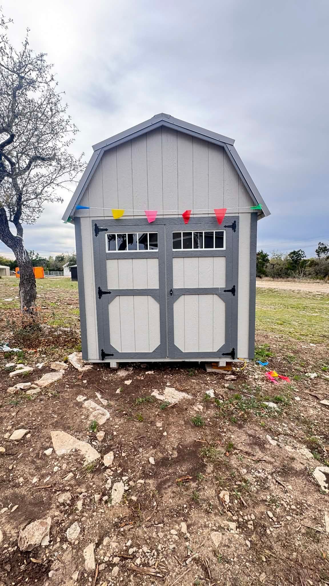 A light-colored storage shed with a gambrel roof and decorative bunting, situated on a rocky, dirt ground.