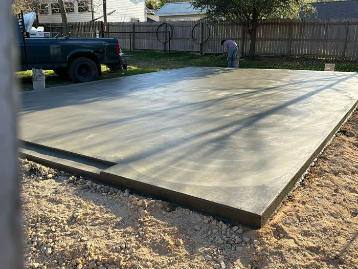 A freshly poured rectangular concrete foundation sits on a construction site under a clear blue sky.