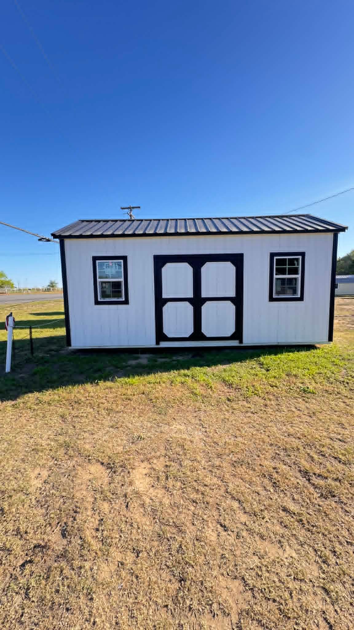 Small white outbuilding with black trim and double doors on a gravel lot under a clear blue sky