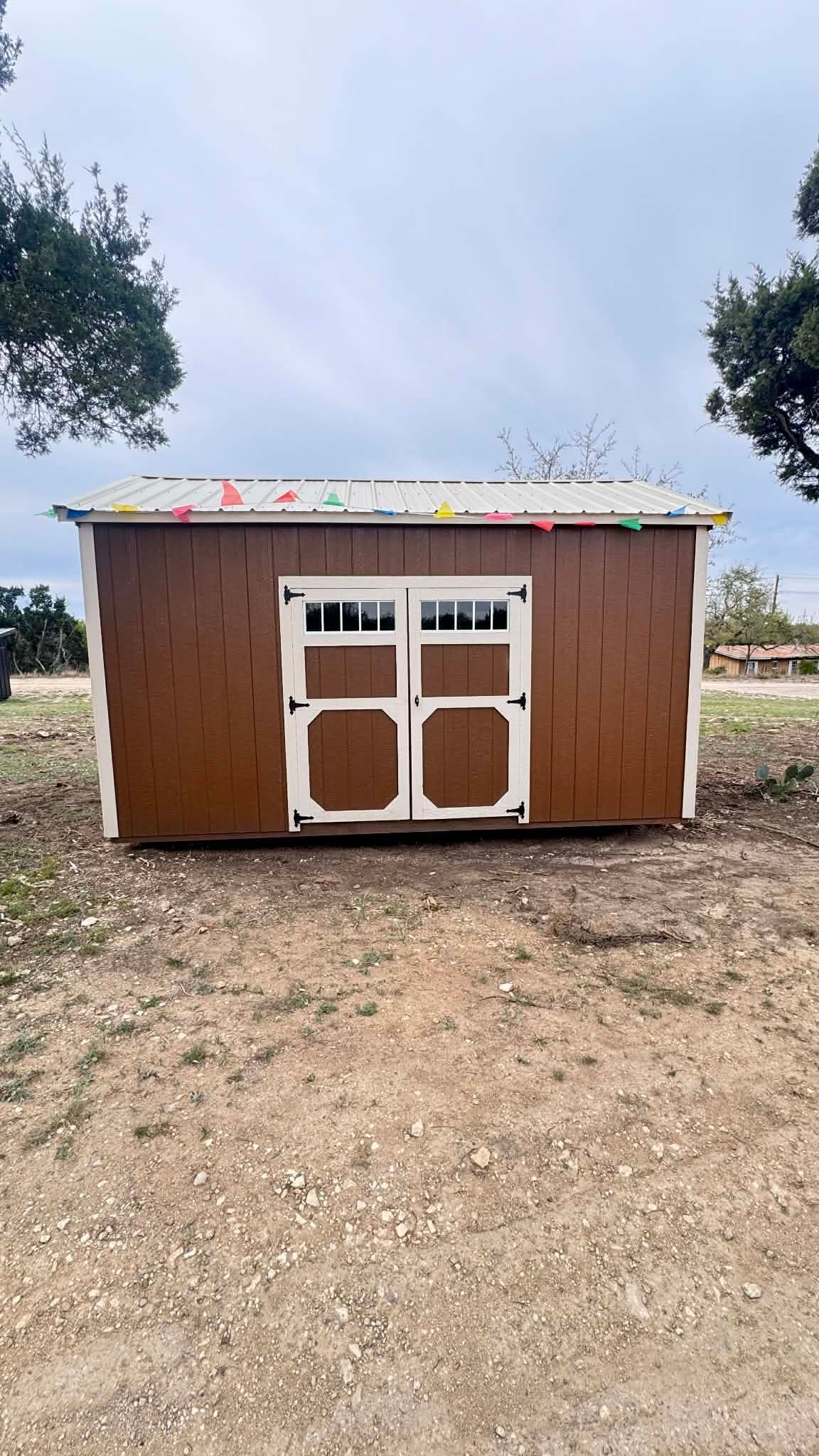 A brown wooden storage shed with white-trimmed double doors, positioned on a gravel lot under a cloudy sky.