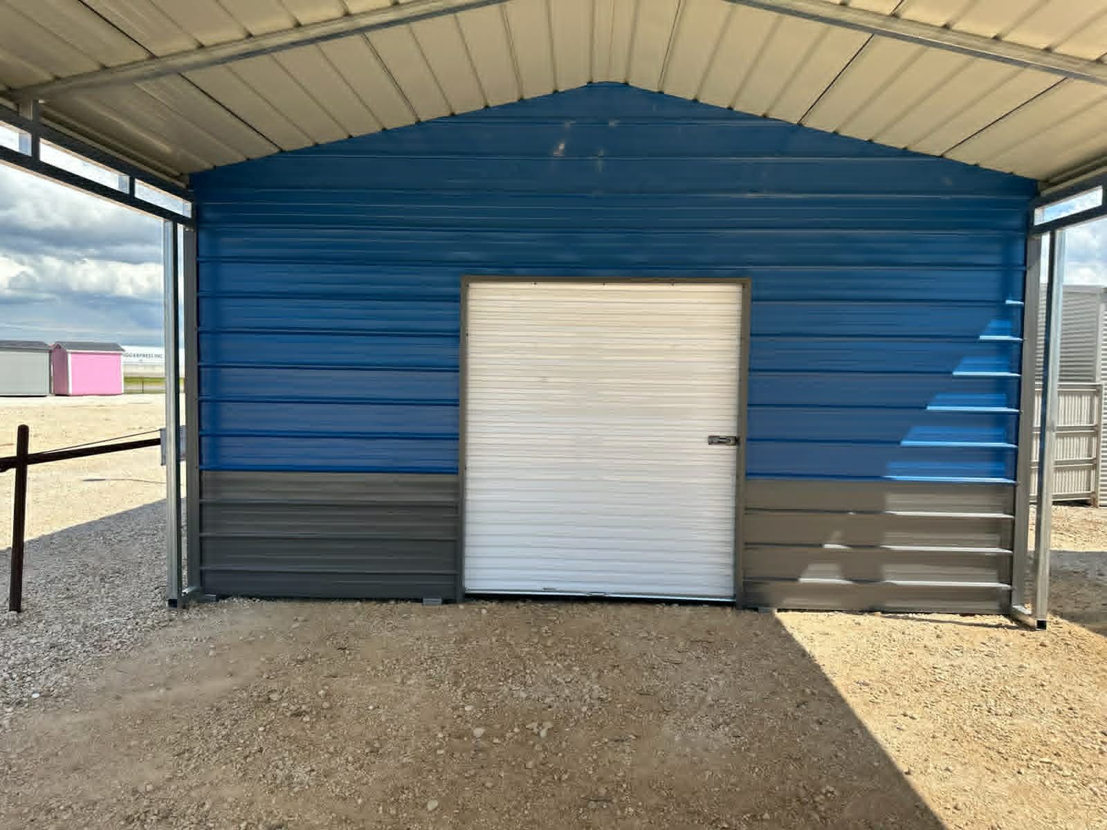 Blue metal shed with a white door under a covered roof on a gravel lot