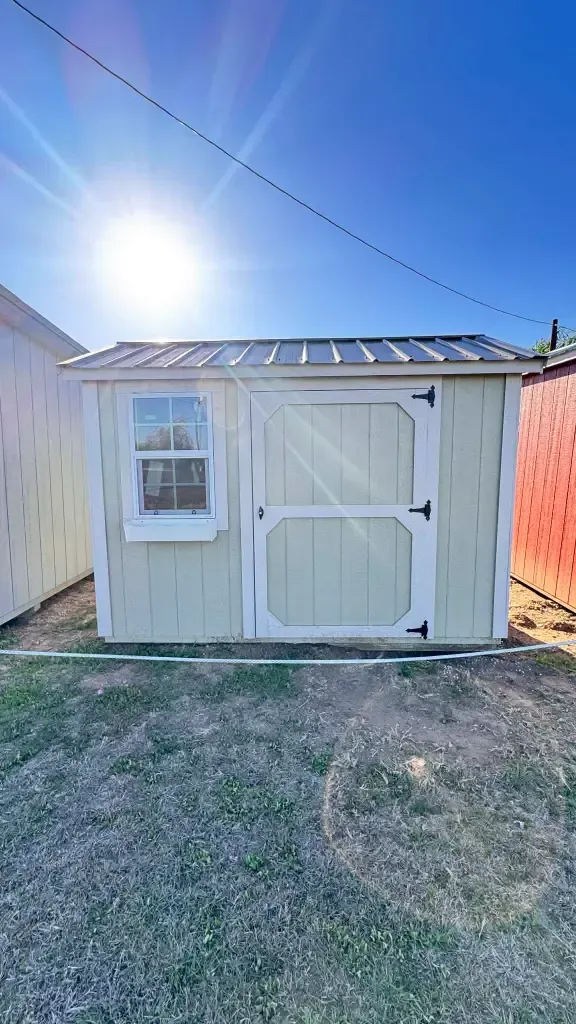 Small beige shed with white double doors and a window in a sunny backyard.