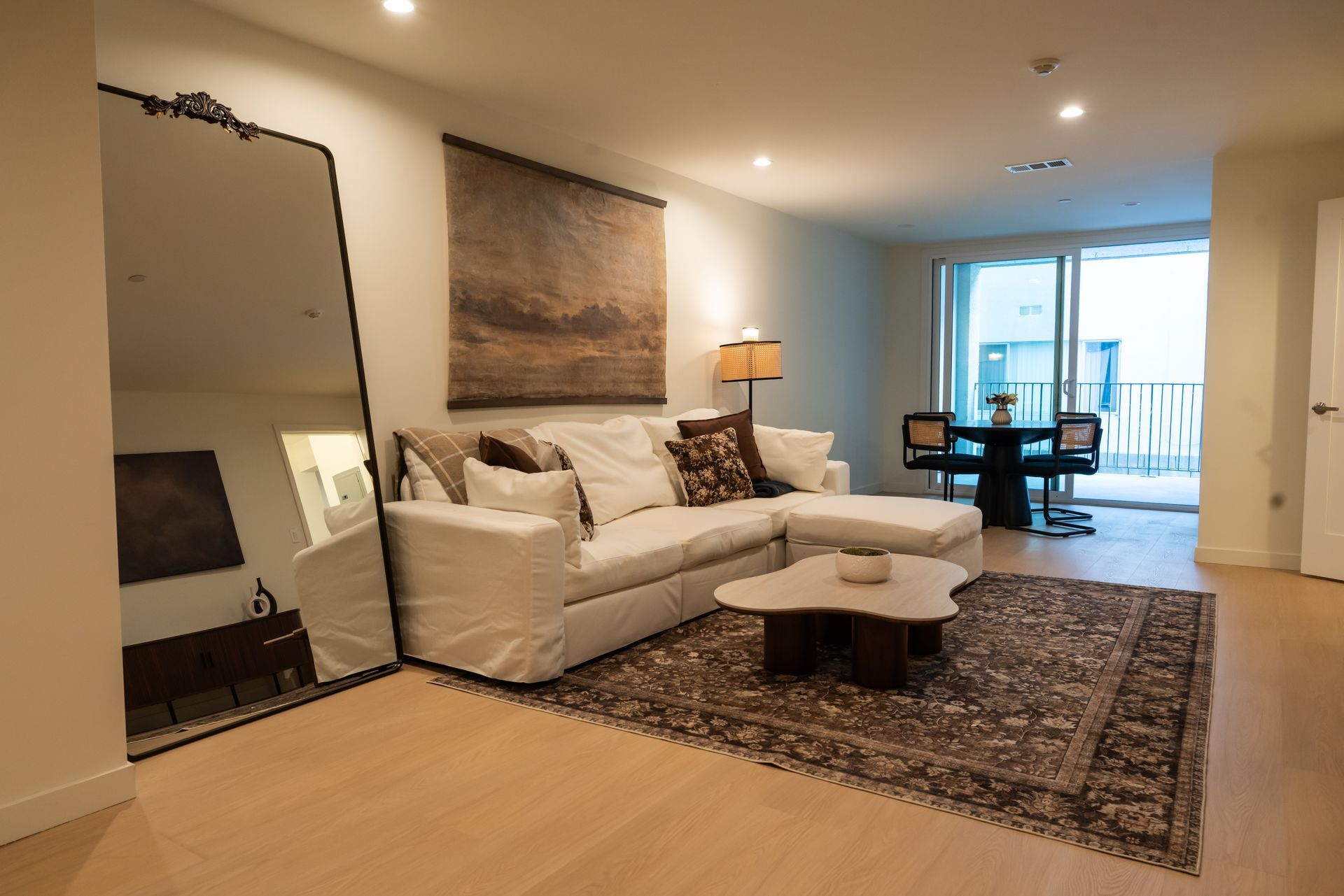 Living room with white sofa, large mirror, rug, and dining area leading to a balcony.