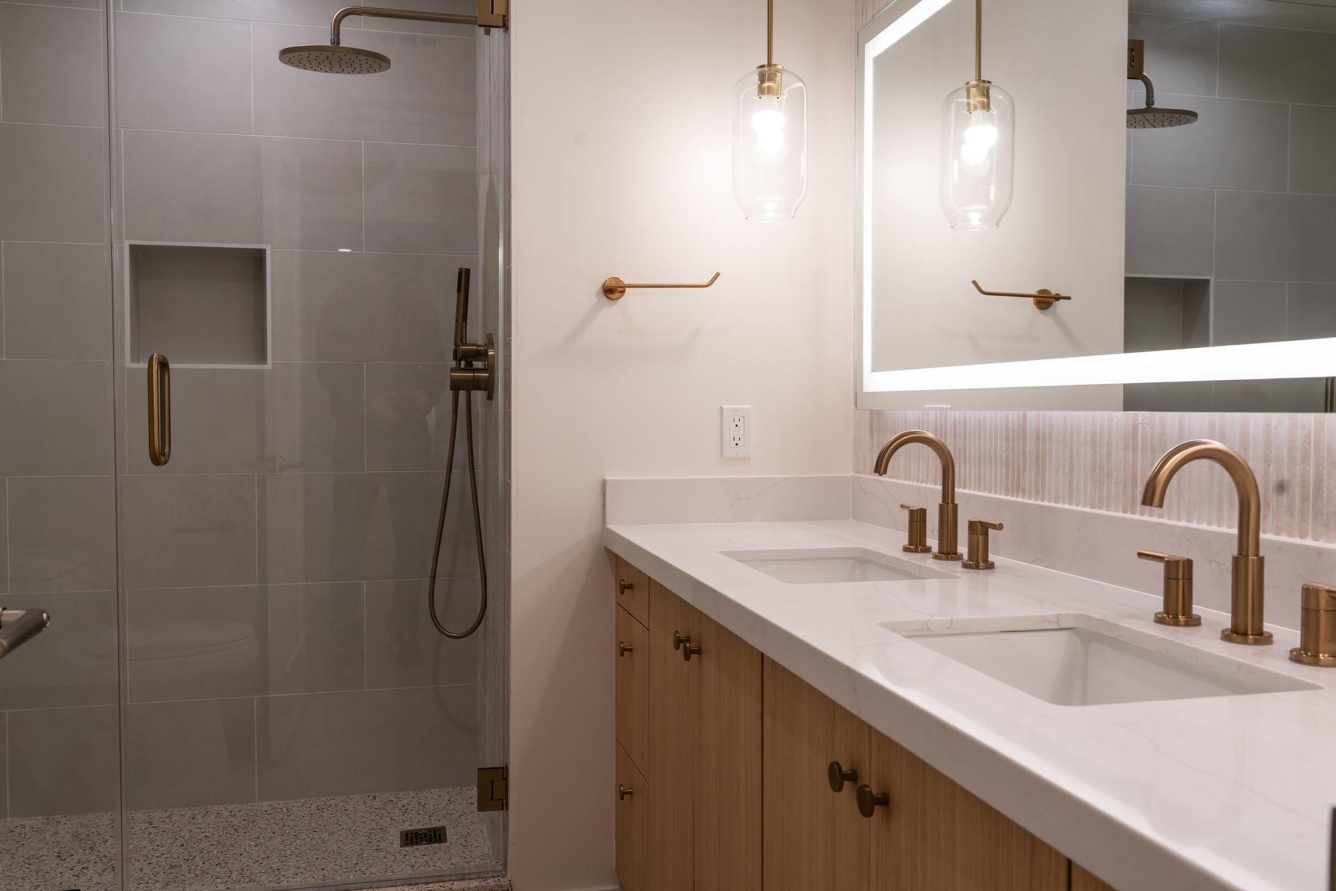 Modern bathroom with glass shower, double sinks, and wood cabinetry. Gold fixtures and recessed lighting.