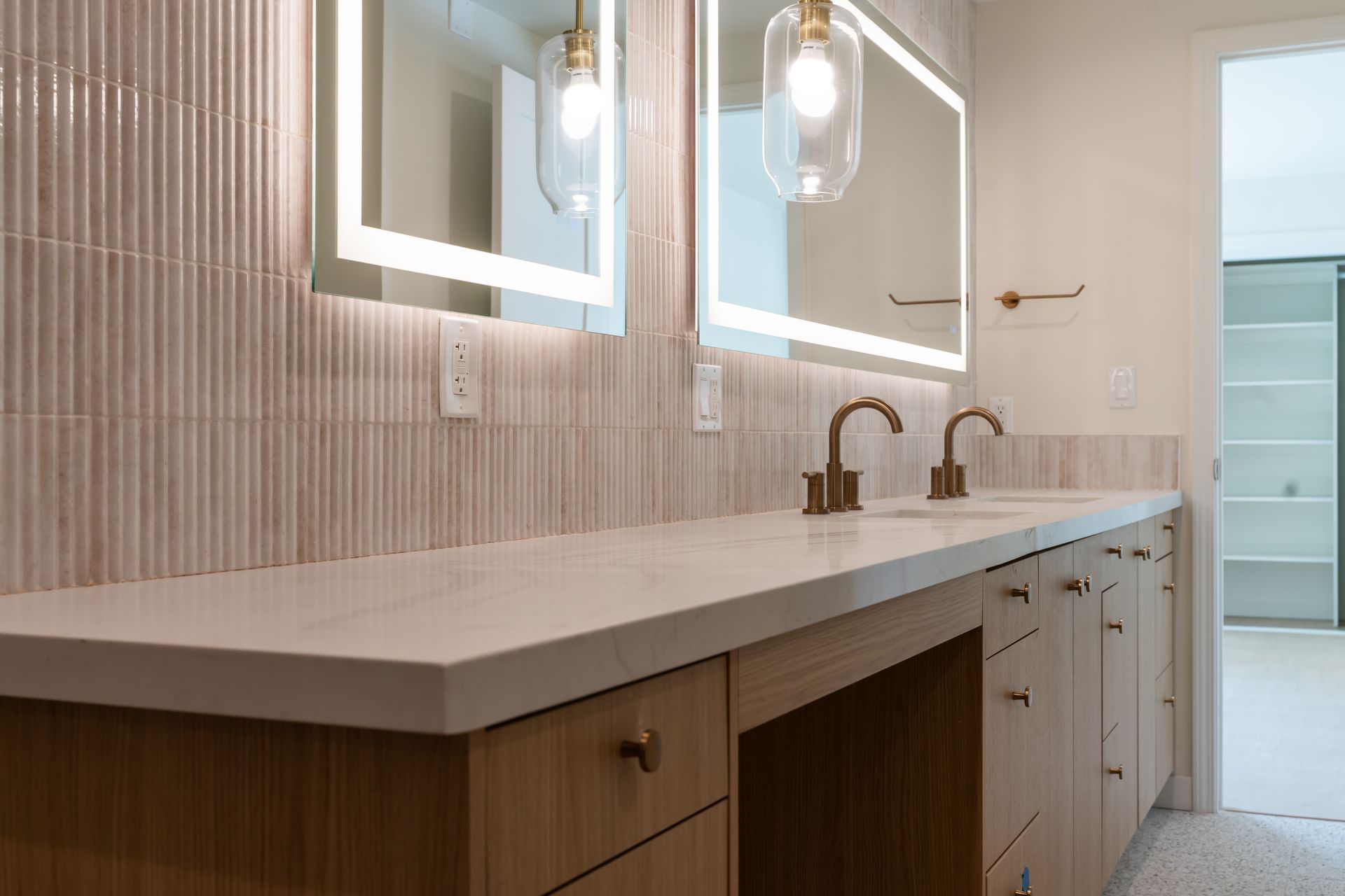 Bathroom with light-colored vanity, two illuminated mirrors, gold fixtures, and textured wall tiles.