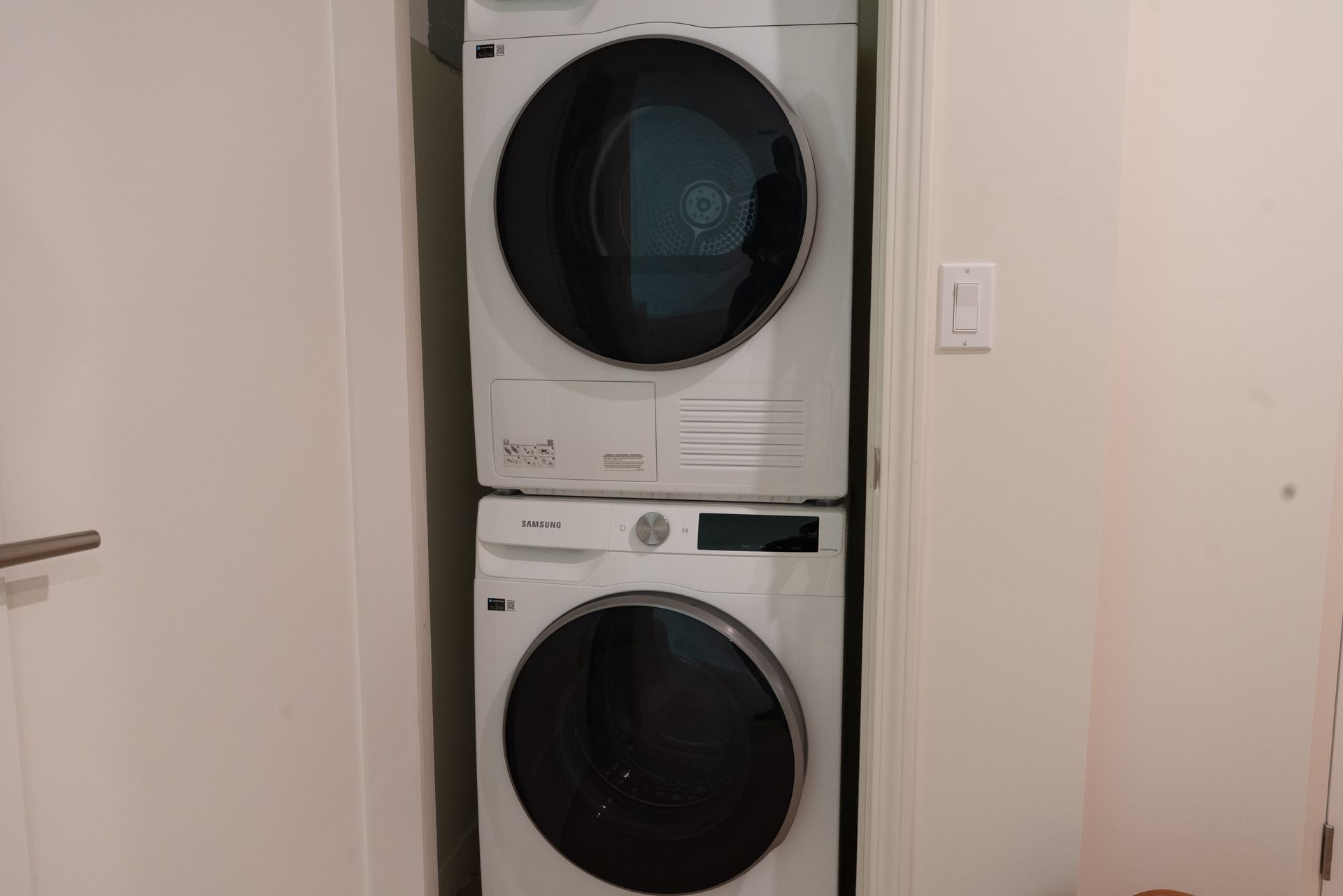 Stacked white washing machine and dryer in a narrow closet with a light switch.