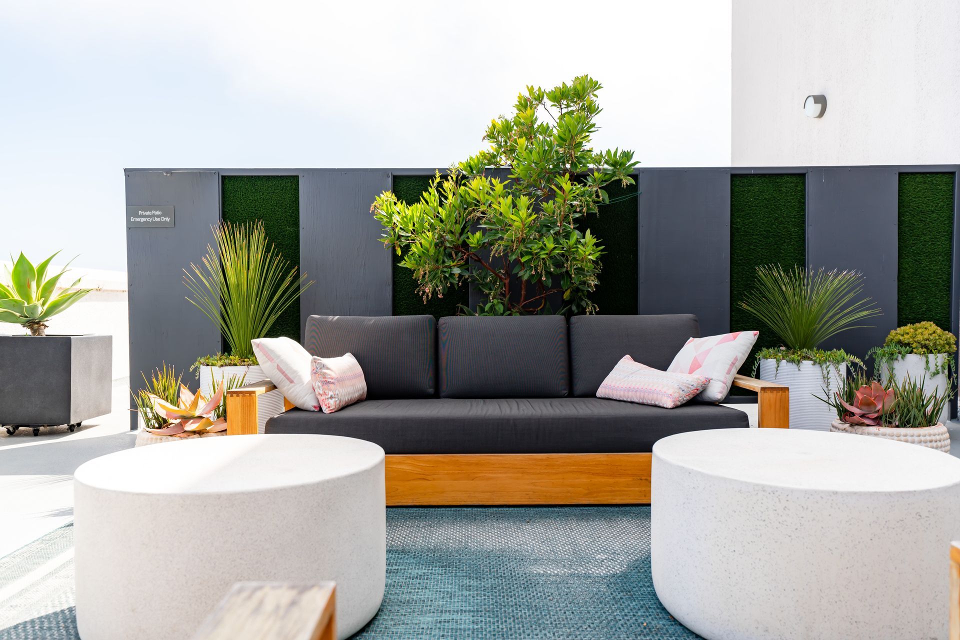 Outdoor seating area with a dark gray couch, round white ottomans, and lush greenery.