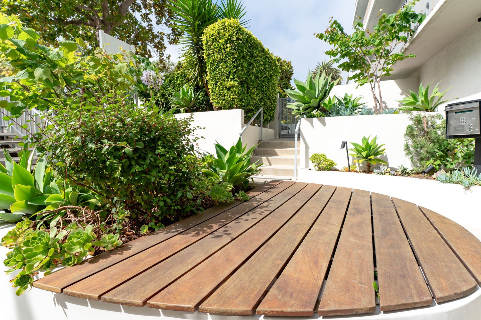 Wooden deck with greenery, stairs in the background, between white walls.