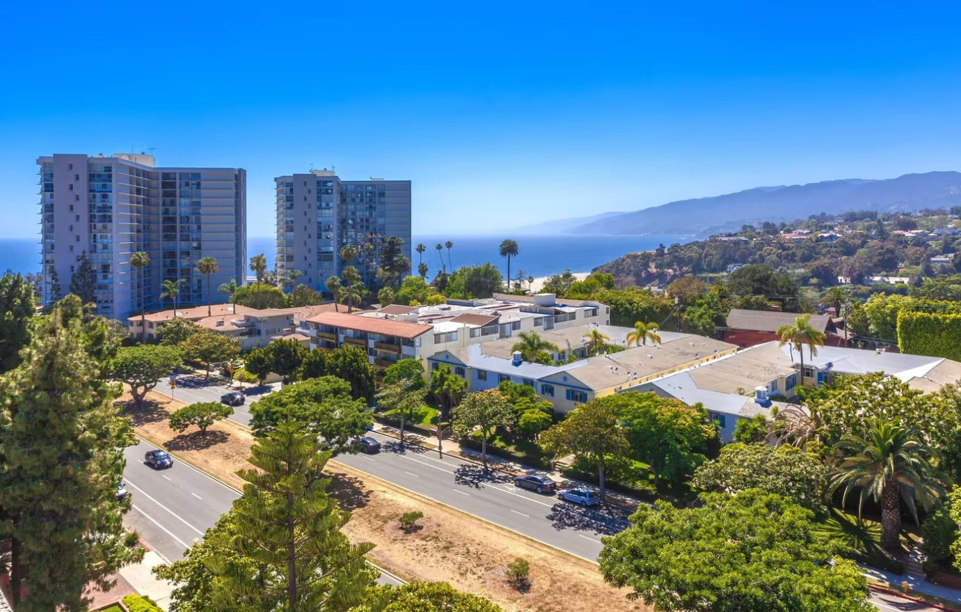 View of buildings, road, and ocean on a sunny day. Blue sky, green trees, and white buildings.
