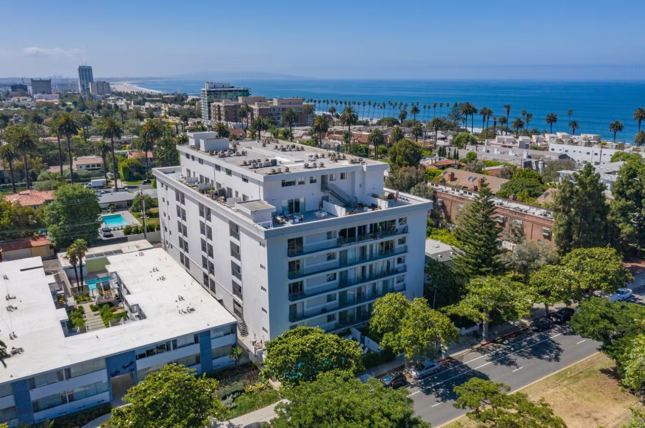 Aerial view of a white apartment building near the ocean in a sunny environment with palm trees.